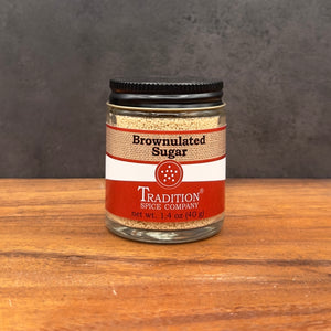 A glass jar of Tradition Spice Company Brownulated Sugar with a black lid, set on a wooden surface against a dark gray background.