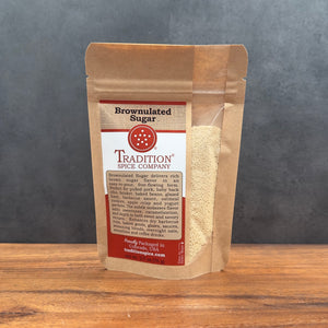 A stand-up pouch of Tradition Spice Company Brownulated Sugar standing on a wooden surface against a dark gray background. The package is made of brown kraft paper with a clear window showing the light tan sugar inside, and a white and red label on the front.