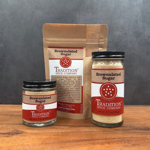 Three jars and one standing pouch of brownulated sugar from Tradition Spice Company arranged on a wooden surface against a dark gray backdrop.