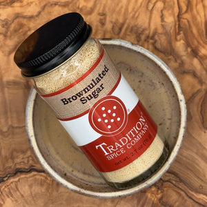 A glass jar of Tradition Spice Company Brownnulated Sugar with a black lid, resting on a rustic ceramic dish over a textured wood background.