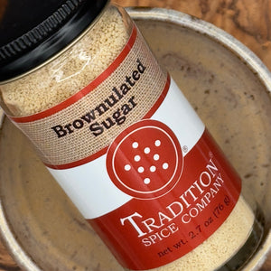 A close-up of a glass jar of Tradition Spice Company Brownulated Sugar, showing the light tan granulated sugar inside, a red and white label, and a black lid, set inside a rustic metal bowl.
