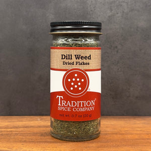 A glass jar of Tradition Spice Company Dill Weed Dried Flakes, displayed on a wooden surface against a dark gray textured background.
