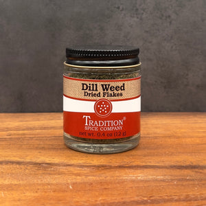 A glass jar of Tradition Spice Company Dill Weed Dried Flakes with a black lid, set on a wooden surface against a dark gray background.