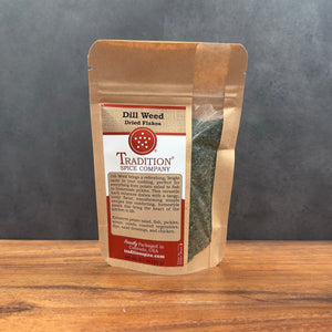 A stand-up pouch of Tradition Spice Company Dill Weed Dried Flakes standing on a wooden surface against a dark gray background.