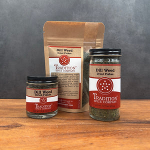Three spice products from Tradition Spice Company on a wooden surface against a dark gray background: a small glass jar, a medium glass jar, and a brown paper pouch, all labeled Dill Weed Dried Flakes.