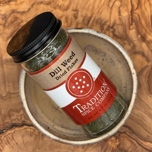 A glass jar of Tradition Spice Company Dill Weed Dried Flakes spice, featuring a black lid and a red and white label, resting inside a rustic ceramic bowl on a textured wood background.