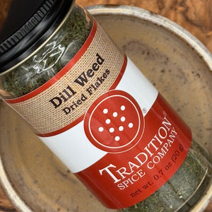A close-up of a glass jar of Tradition Spice Company Dill Weed Dried Flakes, showing the green dried herbs inside and the red, white, and beige label, resting on a rustic metal plate.