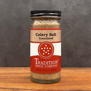 A glass jar of Tradition Spice Company Celery Salt Granulated, featuring a black metal lid and a label with red, white, and burlap textures, set on a wooden surface against a dark gray background.