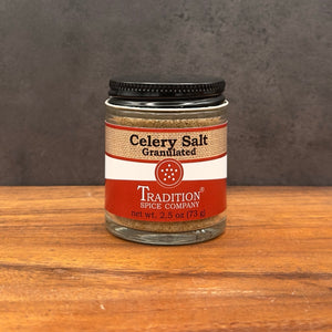 A jar of Tradition Spice Company Celery Salt Granulated, sitting on a wooden surface against a dark gray textured background.