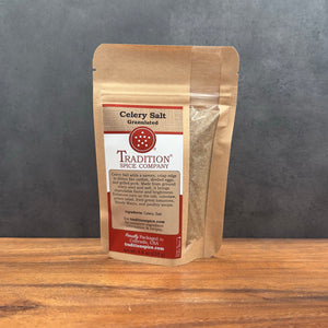 A stand-up kraft paper pouch of Tradition Spice Company Celery Salt, Granulated, standing on a wooden surface against a dark gray background.