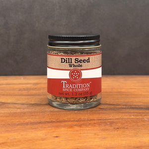 A clear glass jar of Whole Dill Seed from Tradition Spice Company rests on a wooden surface, featuring a black lid and red-beige label—ideal as a pickling spice for your favorite recipes.