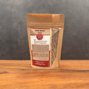 A brown paper pouch of Whole Dill Seed from Tradition Spice Company stands on a wooden surface, featuring a red and white label listing uses and ingredients, set against a gray textured background.