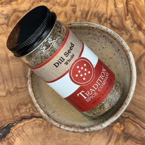 A glass jar labeled Whole Dill Seed rests at an angle in a speckled ceramic bowl on a wooden surface. The jar contains light brown whole dill seeds, ideal as a pickling spice or for seasoning various dishes.