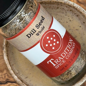 A glass jar labeled Whole Dill Seed rests on a speckled ceramic bowl. The jar displays dried dill seeds, commonly used in pickling, and features a red and beige label.