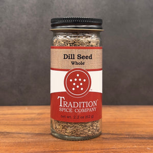 A 2.2 oz (62 g) glass jar of Whole Dill Seed from Tradition Spice Company sits on wood with a gray background. It features a red and beige label with bold text and a circular design, perfect for pickling.