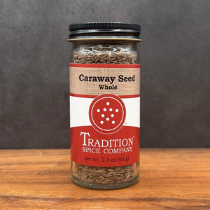 A glass jar of Whole Caraway Seed from Tradition Spice Company rests on a wooden surface, featuring a black lid and a red and white label with the product name and weight.