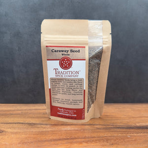 A brown resealable pouch of Whole Caraway Seed sits on a wooden surface, with a clear window showing the seeds and a red and white label featuring product details.