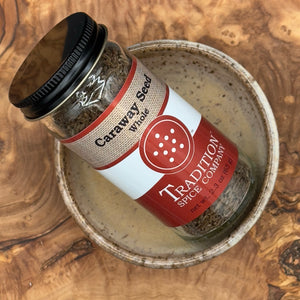 A jar of Whole Caraway Seed from Tradition Spice Company, with a red and white label and black lid, sits in a ceramic bowl on a wood-grain surface, highlighting the quality of these aromatic seeds.