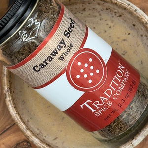 A jar of Whole Caraway Seed from Tradition Spice Company sits in a speckled ceramic bowl, featuring a red and beige label with a net weight of 2.3 oz (65 g).