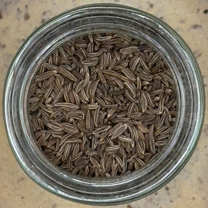 A glass jar of Tradition Spice Company's Whole Caraway Seed, containing brown, oval seeds, viewed from above on a beige surface.