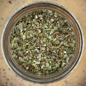 Top-down view of dried chopped herbs or spices in a clear glass jar with a metal rim, set on a tan speckled surface.