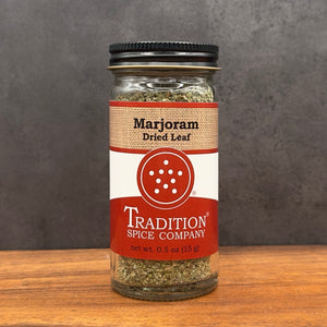 A glass jar of Tradition Spice Company Marjoram Dried Leaf on a wooden surface against a dark gray textured background.
