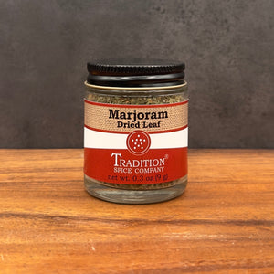 A glass jar of Tradition Spice Company Marjoram Dried Leaf on a wooden surface against a dark gray background.