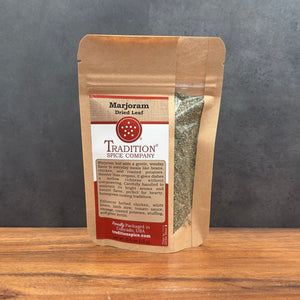 A stand-up kraft paper pouch of Tradition Spice Company Marjoram Dried Leaf, standing on a wooden surface against a dark gray background.