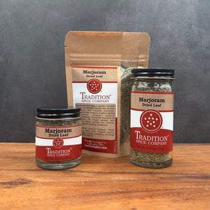 Three jars and one standing pouch of Tradition Spice Company Marjoram Dried Leaf on a wooden surface against a dark gray background.