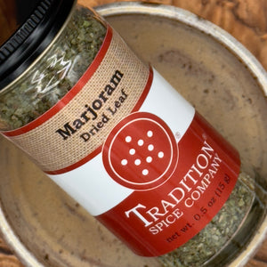 A close-up of a jar of Tradition Spice Company Marjoram Dried Leaf spice, resting inside a rustic metal bowl.