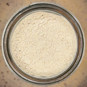 A top-down view of a light beige powder, likely flour or cosmetic powder, inside a clear glass jar with a silver metal rim.