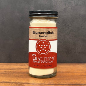 A glass jar of Tradition Spice Company Horseradish Powder with a black metal lid, set on a wooden surface against a dark gray background.