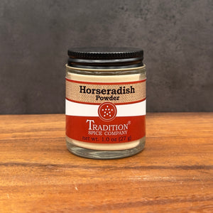 A glass jar of Tradition Spice Company Horseradish Powder with a black lid, set on a wooden surface against a dark gray background.