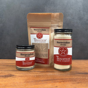 Three spice product containers from Tradition Spice Company on a wooden surface against a dark gray background: a small glass jar, a large glass jar, and a brown paper pouch, all labeled Horseradish Powder.