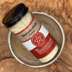 A glass jar of Tradition Spice Company Horseradish Powder with a black lid and red-and-white label, resting inside a rustic ceramic bowl on a wood-grain surface.