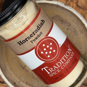 A close-up of a glass jar of Tradition Spice Company Horseradish Powder, showing the cream-colored powder inside and the red, white, and burlap label, resting inside a rustic metal bowl.