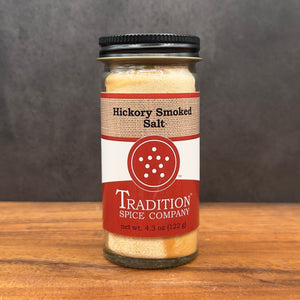 A 4.3 oz (122 g) glass jar of Hickory Smoked Salt sits on a wooden surface, its red and white label displayed. Add bold, wood-fired smokehouse flavor to your next smoky Bloody Mary with this savory seasoning.