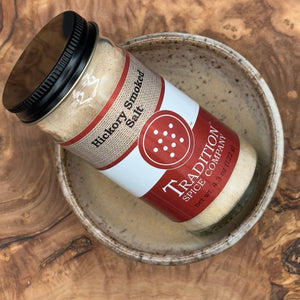 A jar of Hickory Smoked Salt sits in a speckled ceramic bowl on wood. The jar features a black lid and a red and white label.