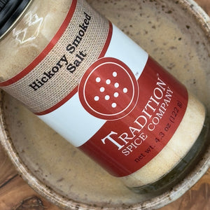 A jar of Hickory Smoked Salt, inspired by wood-fired smokehouses, sits in a ceramic bowl. Its label shows the product name and net weight (4.3 oz/122 g). Ideal for adding rich smoky flavor to cocktails like a Bloody Mary.