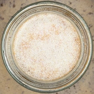 A glass jar of Hickory Smoked Salt, finely ground and light tan in color, seen from above on a beige background—ideal for infusing rich, smoky flavor into your next Bloody Mary or favorite dish.