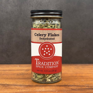 A glass jar of Dehydrated Celery Flakes sits on a wooden surface against a dark background, featuring a red and beige label that emphasizes the garden-fresh taste of this handy seasoning.