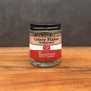 A glass jar of Dehydrated Celery Flakes with a black lid and red-beige label sits on wood, against a dark background, offering vibrant garden-fresh flavor in every sprinkle.