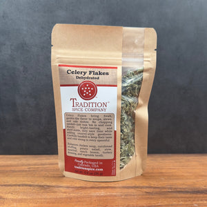 A brown resealable pouch of Dehydrated Celery Flakes stands on a wooden surface. A clear window displays the dried green flakes, ideal for adding garden-fresh celery flavor to dishes. The background is dark gray stone.