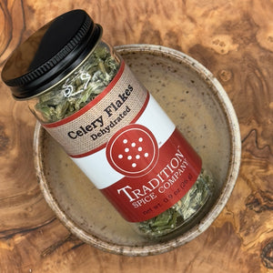 A jar labeled Dehydrated Celery Flakes rests at an angle in a speckled ceramic bowl on a wooden surface, offering vibrant garden-fresh flavor and celery seasoning.