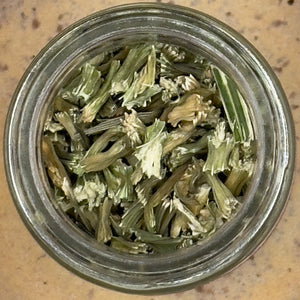 A glass jar of Dehydrated Celery Flakes—dried, chopped green and white stems and leaves—sits on a speckled beige surface, offering garden-fresh celery flavor.