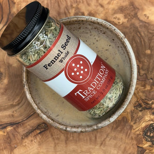 A jar of Whole Fennel Seed sits in a beige ceramic bowl on a wooden surface—great for seasoning Italian sausage or enhancing rustic breads. The jar features a red and beige label with black text and is topped with a black lid.