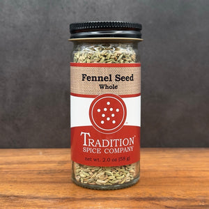 A glass jar of Whole Fennel Seed sits on a wooden surface, with a black lid and red and tan label—ideal for seasoning Italian sausage or baking rustic breads.