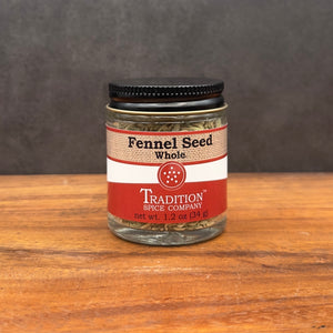 A glass jar of Whole Fennel Seed from Tradition Spice Company rests on wood with a dark backdrop. Ideal for seasoning Italian sausage or breads, its tan and red label shows the product name and 1.2 oz (34 g) net weight.