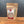 A brown resealable pouch labeled Whole Fennel Seed sits on a wooden surface with a gray background. The clear window displays light green seeds, ideal for rustic breads or seasoning Italian sausage.