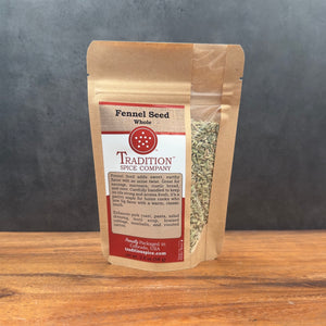 A brown resealable pouch labeled Whole Fennel Seed sits on a wooden surface with a gray background. The clear window displays light green seeds, ideal for rustic breads or seasoning Italian sausage.